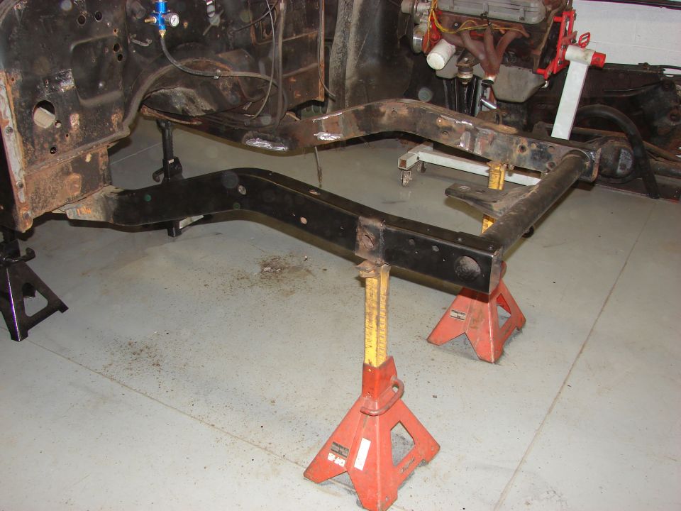 Rusty vehicle frame on red and yellow jack stands with engine visible in background
