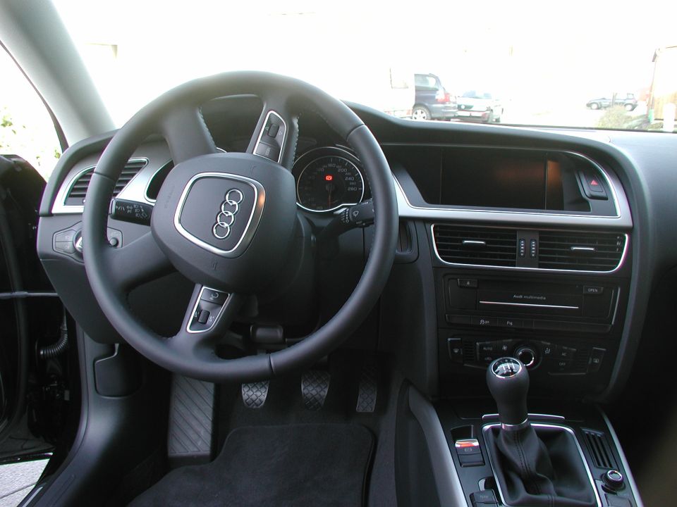 Car interior with steering wheel, dashboard, and gear shift visible. The Audi logo is on the steering wheel