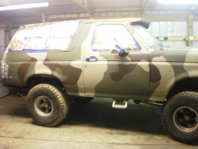 Side view of a camouflage-painted Ford Bronco with masked windows in a shop