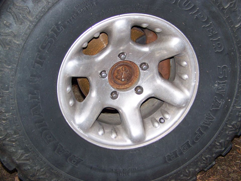 Close-up of a 1992 Jeep YJ wheel with a Super Swamper tire. The wheel is silver with five spokes and visible lug nuts