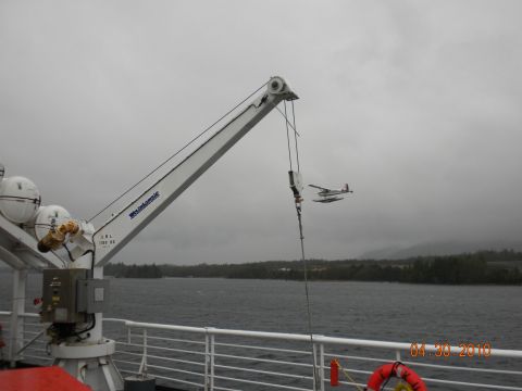 1989 Ford Bronco - A seaplane flies over a body of water near a forested shoreline, with a crane visible on a boat in the foreground.