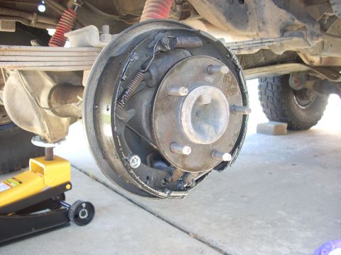 1989 Ford Bronco - Close-up of a rear drum brake assembly, with a yellow hydraulic jack supporting the vehicle.