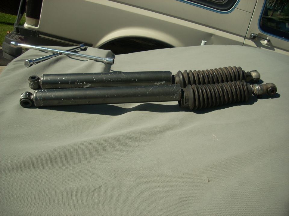 Two black shock absorbers with rubber bellows and a lug wrench on a grey tarp. Part of a white vehicle is visible in the background