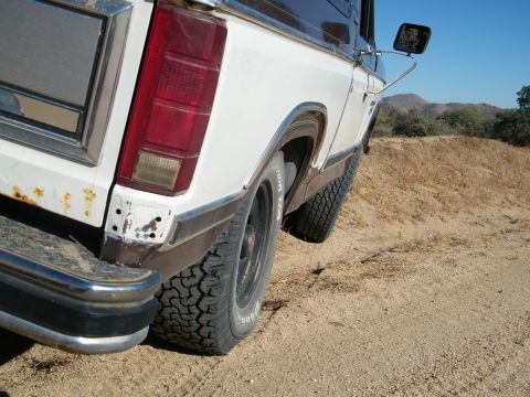 1982 Ford Bronco - This Ford Bronco is shown from the rear quarter angle, driving on a dirt road with dry grass and hills in the background.