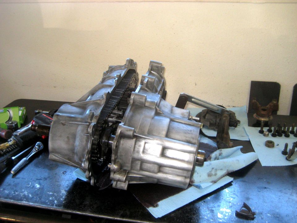 Transfer case assembly with visible chain and gears, resting on a workbench with tools