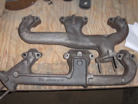 1970 Chevrolet Camaro - Two exhaust manifolds are laid out on a wooden surface, showing GM part numbers and casting marks.