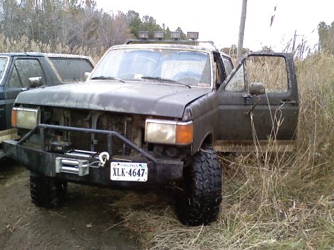 1990 Ford Bronco - A dark gray Ford Bronco sits in tall grass with its driver's door open, modified with a winch and off-road tires.