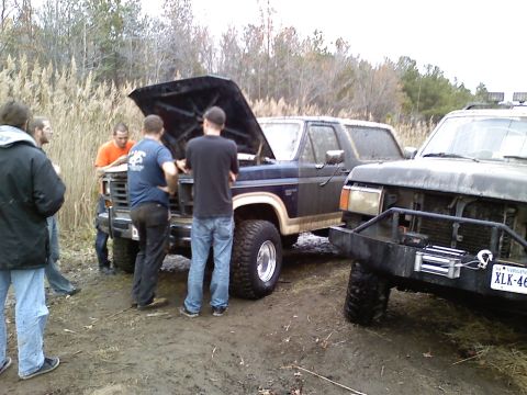 1990 Ford Bronco - Several people gather around a Ford Bronco with its hood open, parked next to another vehicle in a muddy, outdoor setting.