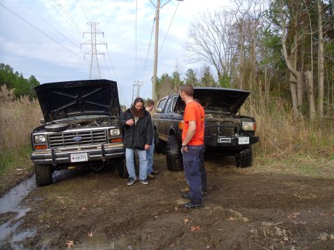 1985 Ford Bronco - Two Ford Broncos with hoods open are stuck in a muddy area with power lines overhead.