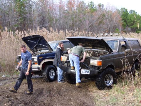 1985 Ford Bronco - Two men work on their off-road vehicles, a dark Bronco and a blue pickup, in a muddy field with dry reeds.