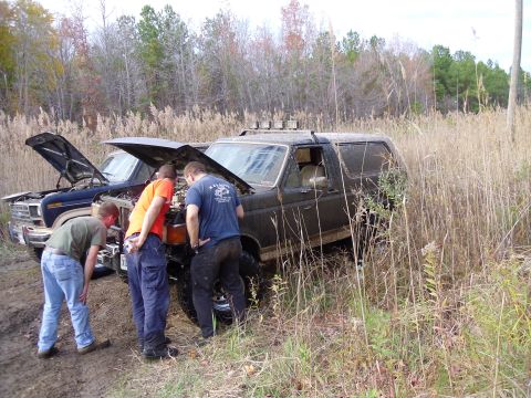 1985 Ford Bronco - Two vehicles are stuck in the mud, with three men looking under the hood of the lead vehicle.