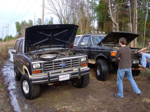 1985 Ford Bronco - Two Ford Broncos, both with hoods open, sit in a muddy, wooded area.