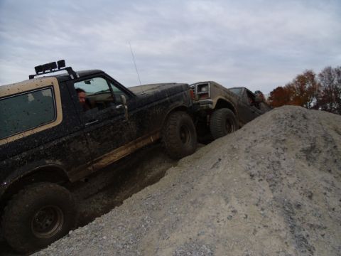 1985 Ford Bronco - Two SUVs navigate a challenging off-road terrain, kicking up dirt as they ascend a steep incline.