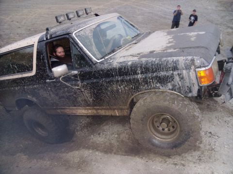 1985 Ford Bronco - A muddy Ford Bronco navigates off-road terrain, with two people watching in the background.
