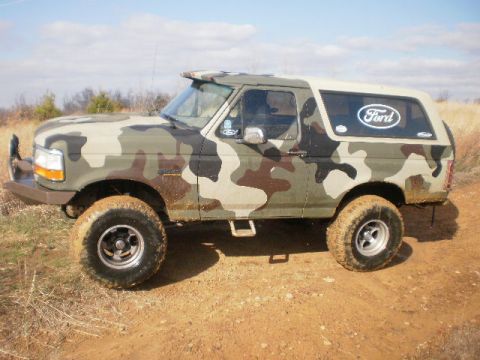 1995 Ford Bronco - A camouflaged Ford Bronco with a tan hardtop sits on a dirt road.