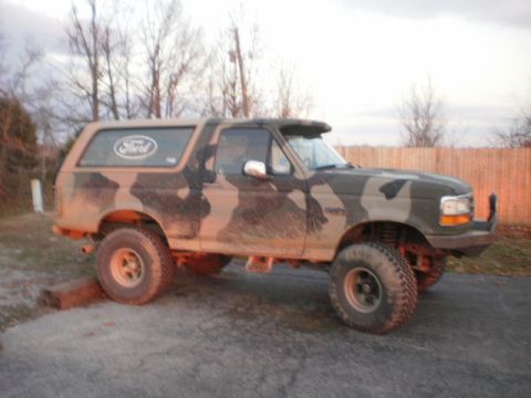 1995 Ford Bronco - This lifted Ford Bronco features a camouflage paint job and is parked on a gravel surface near a wooden fence.