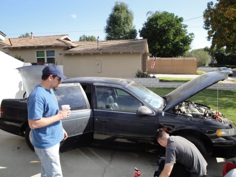 1995 Ford Taurus - Two men work on a black 1995 Ford Taurus sedan parked in a driveway with its hood and doors open.