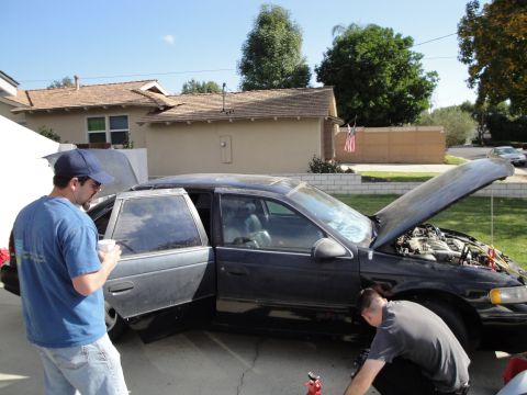 1995 Ford Taurus - Two people work on a dark sedan with its hood and doors open, revealing the engine and interior.