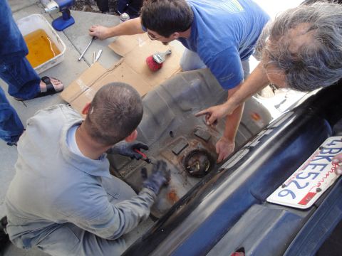 1995 Ford Taurus - Several people are gathered around a car, working on its fuel tank which has been removed from the vehicle.