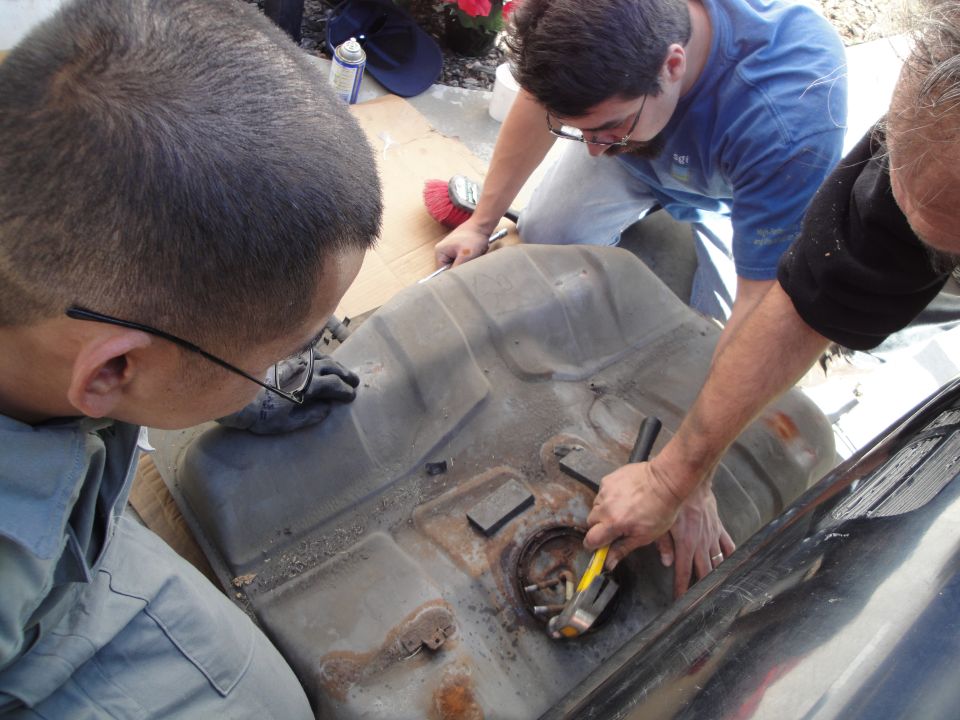 Three people gathered around a rusty, removed fuel tank, one using a hammer and tool on the fuel pump assembly