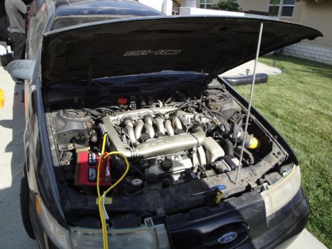 1995 Ford Taurus - The engine bay of a black 1995 Ford Taurus SHO is visible with the hood raised, revealing the V8 engine and a red battery.