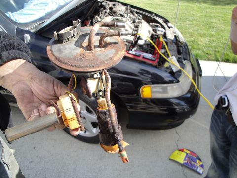 1995 Ford Taurus - A mechanic holds a rusty fuel pump assembly in front of a black 1995 Ford Taurus with its hood open and jumper cables attached.