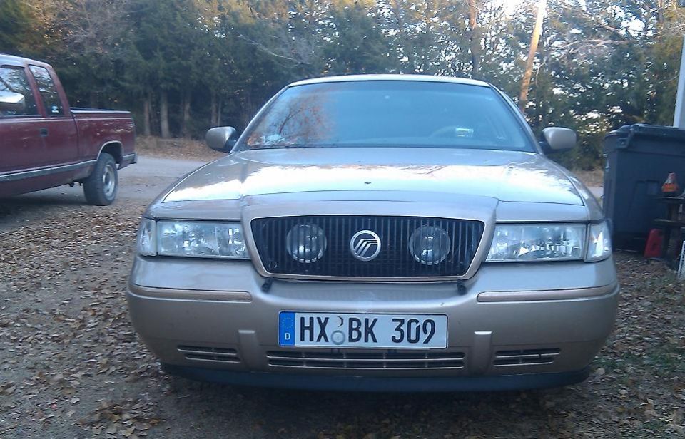 Front view of a gold 2000 Mercury Grand Marquis with a black grille and a German license plate. A red pickup truck is parked to the left