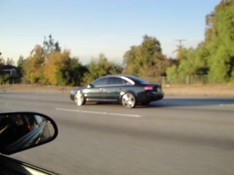 1995 Ford Taurus - A dark sedan is captured in profile while driving on a highway, its wheels appearing to be aftermarket modifications.