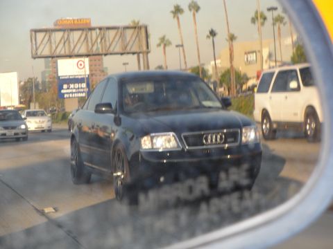 1995 Ford Taurus - A black Audi sedan is seen on the highway, reflected in a side mirror, with other vehicles and palm trees in the background.