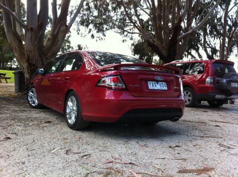 2010 Ford Falcon - Two red vehicles are parked outdoors on a gravel surface near trees. One is a Ford Falcon sedan, the other is an SUV.