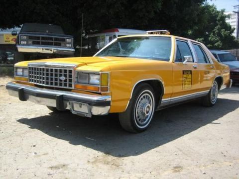 1991 Ford Crown Victoria - A yellow taxi cab with a chrome grille and bumper is parked outdoors, with other vehicles in the background.