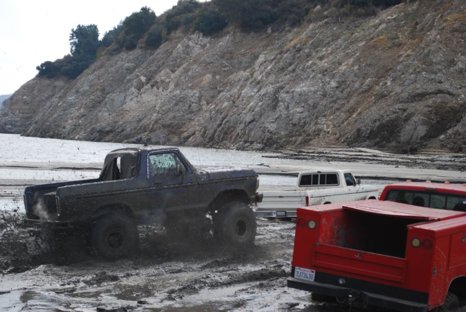 Muddy trucks driving through a wet, rocky area with a large hill in the background