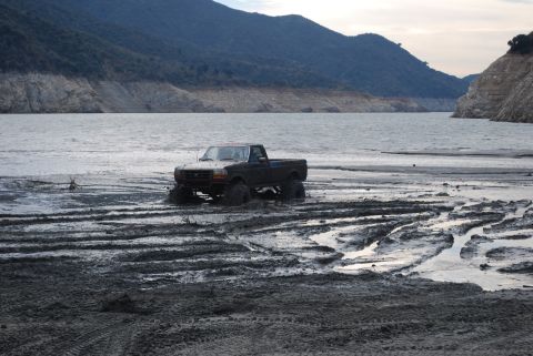 1996 Ford Bronco - A lifted Ford F-Series truck is bogged down in thick mud by a large lake with dry, mountainous terrain in the background.