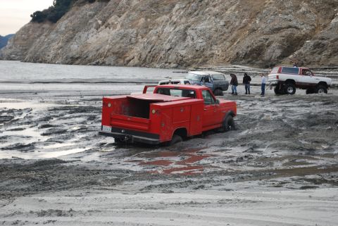 1996 Ford Bronco - Several vehicles are stuck in the mud near a rocky shoreline, with a red pickup truck prominently featured.