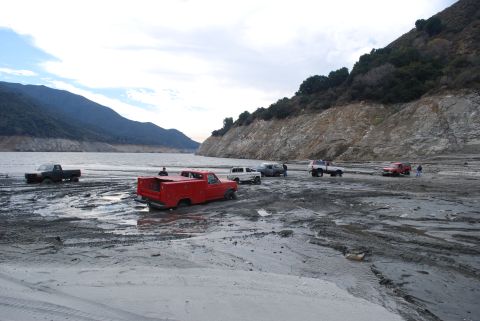 1996 Ford Bronco - Multiple trucks are stuck in the mud near a lake, with a steep, rocky incline in the background.