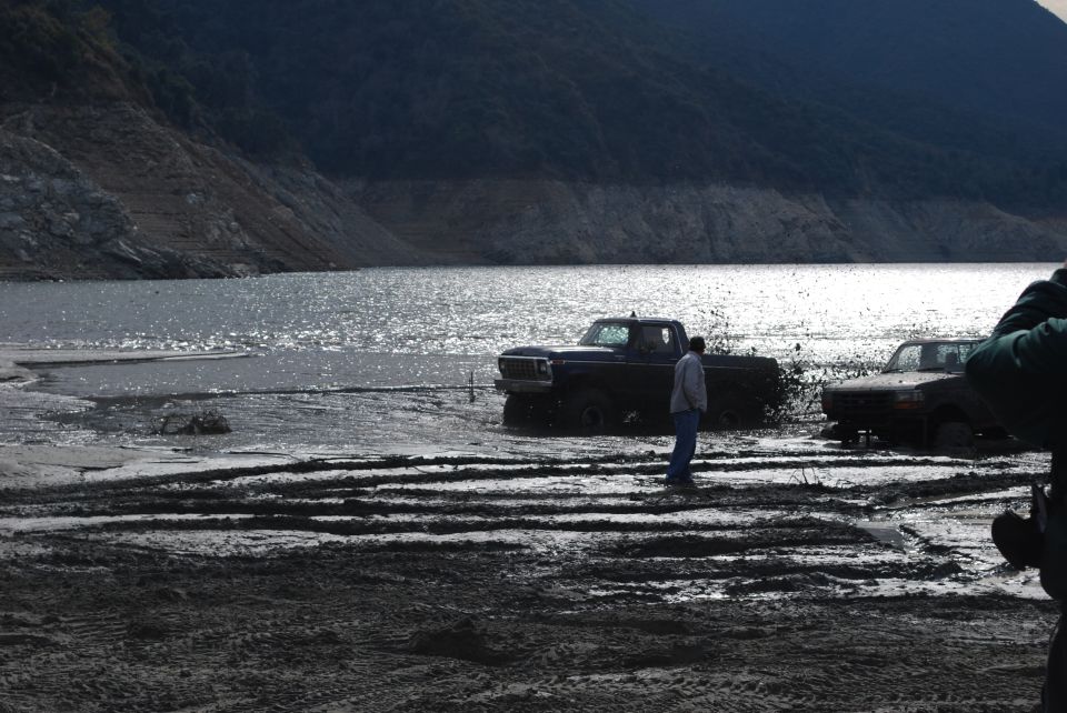 Dark purple truck stuck in mud, facing left, with a person standing nearby. Another truck is visible to the right