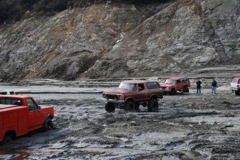 1996 Ford Bronco - Several off-road vehicles navigate a muddy terrain with a rocky hillside in the background.