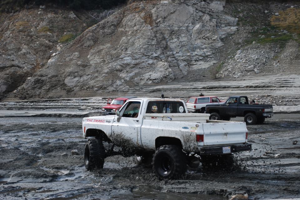 Several lifted trucks driving through mud, one white pickup in foreground