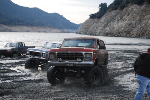 1996 Ford Bronco - Several lifted trucks and SUVs navigate a muddy terrain by a large body of water.