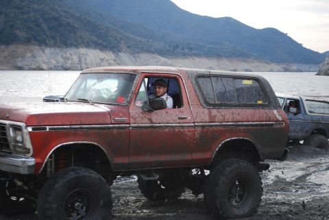1996 Ford Bronco - A muddy red Ford Bronco with a lifted suspension is parked near a body of water alongside another vehicle.