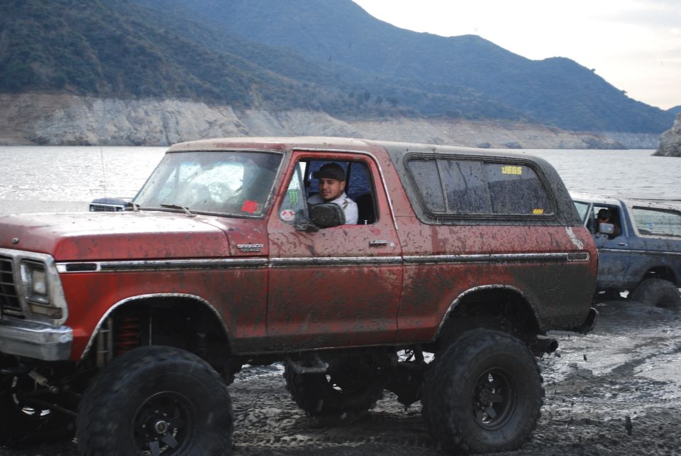 Red Ford Bronco with lifted suspension and large tires, covered in mud, parked next to another vehicle by a body of water
