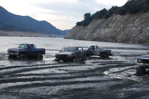 1996 Ford Bronco - Multiple off-road trucks are stuck in thick mud by a large body of water with hills in the background.