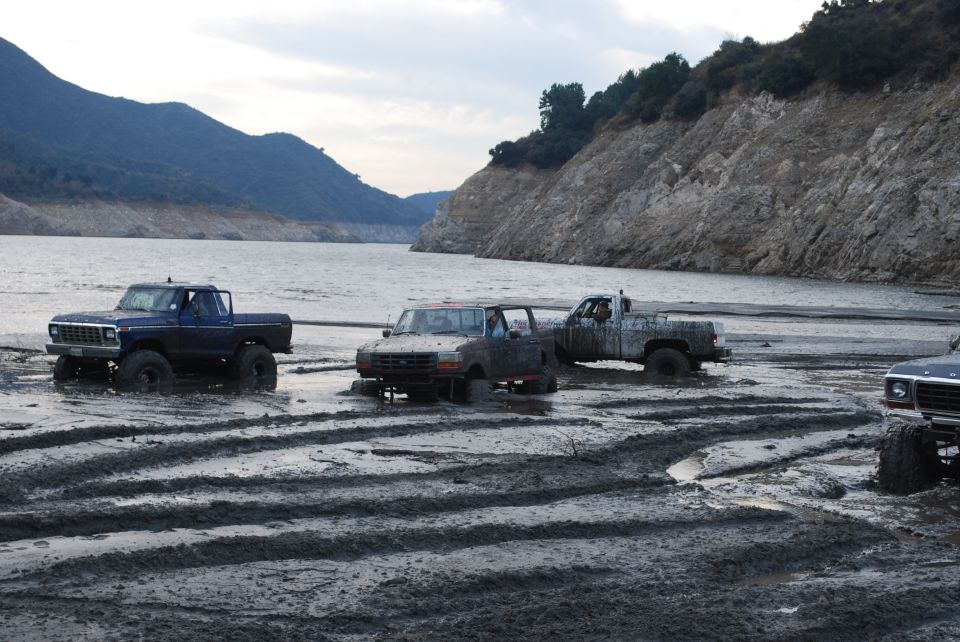 Four-wheel drive trucks stuck in mud near a lake, one blue pickup in foreground, others further back