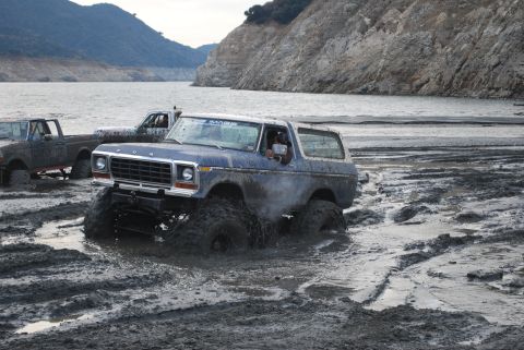1996 Ford Bronco - Multiple off-road vehicles navigate a muddy terrain near a body of water and rocky hills.