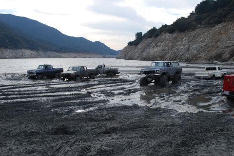 1996 Ford Bronco - Several trucks and a Ford Bronco are stuck in the mud by a body of water.