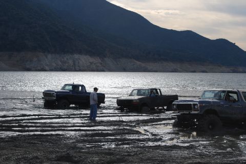 1996 Ford Bronco - Several trucks are stuck in the mud near a large body of water, with a mountain range in the background.