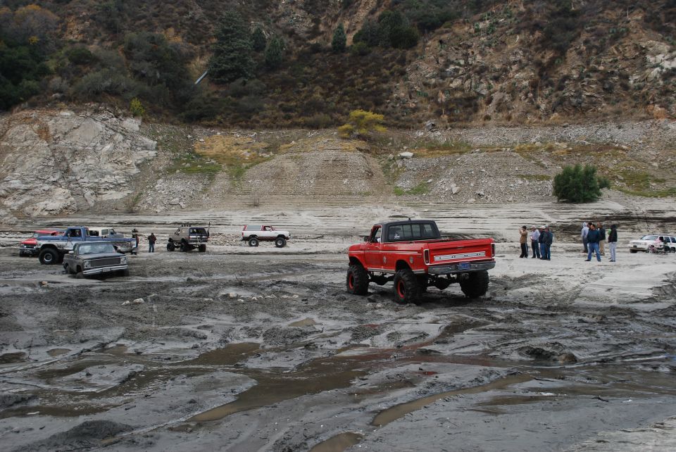 Red pickup truck with lifted suspension and large tires in a muddy area. Other trucks and SUVs are visible in the background