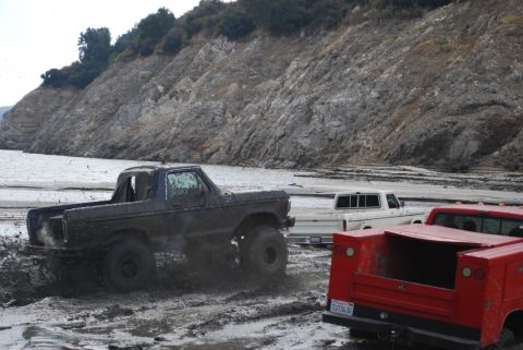 1996 Ford Bronco - Several trucks navigate a muddy terrain with a rocky hillside in the background.