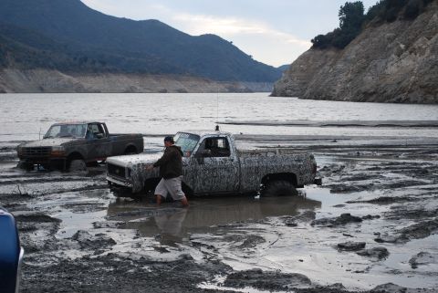 1996 Ford Bronco - Two trucks are stuck in mud near a large body of water with hills in the background.