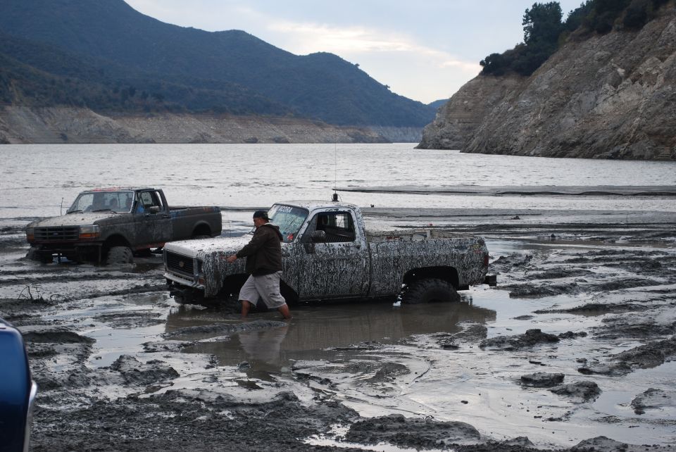 Two trucks stuck in mud with a person pushing one. Water and hills in the background